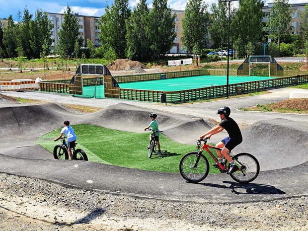 Kolme pyöräilijää ajaa pumptrack-radalla Halkolan liikuntapuistossa, taustalla näkyy monitoimikenttä ja ympäröivä puistoalue.