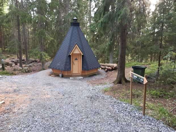 Kaitajärven kota metsäisessä ympäristössä, hirsirakenteinen ja tummalla katolla varustettu tulipaikka, edessä sorapolku ja vieressä opastekyltti sekä roskis