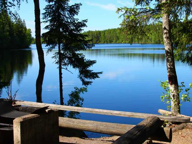 Nuotiopaikka järven rannalla, edessä puisia penkkejä ja tulipaikka. Taustalla kirkas sininen järvi ja vihreä metsä, aurinko valaisee maisemaa.
