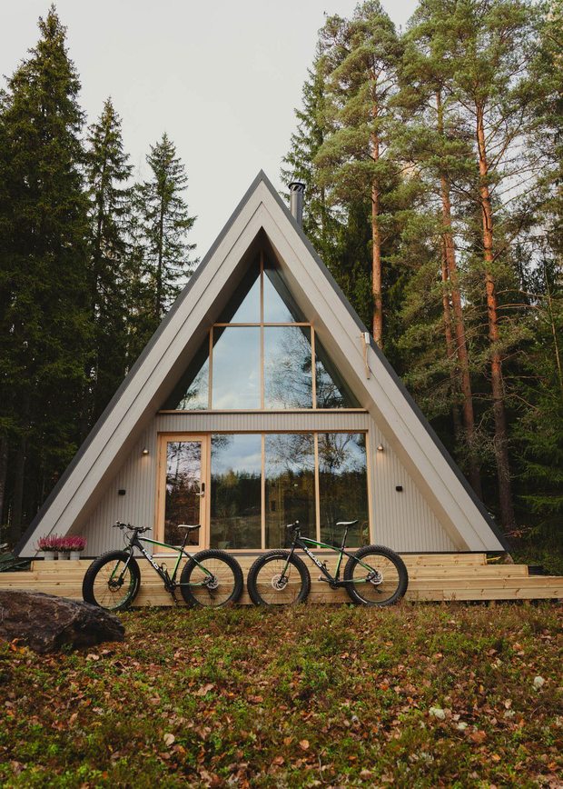 A-muotoinen mökki metsäisessä ympäristössä; edessä puinen terassi ja kolme maastopyörää, taustalla korkeita havupuita