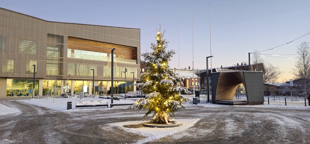 A snow-covered town square at dusk with a decorated Christmas tree glowing with warm lights in the center. Behind it stands a modern building with large glass windows, and on the right a curved wooden pavilion. The ground and trees are covered in snow, creating a calm winter atmosphere.