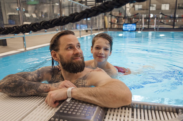 Aikuinen mies ja lapsi hymyilevät uima-altaassa, nojaten altaan reunaan. Taustalla näkyy sisäuima-altaan ympäristö