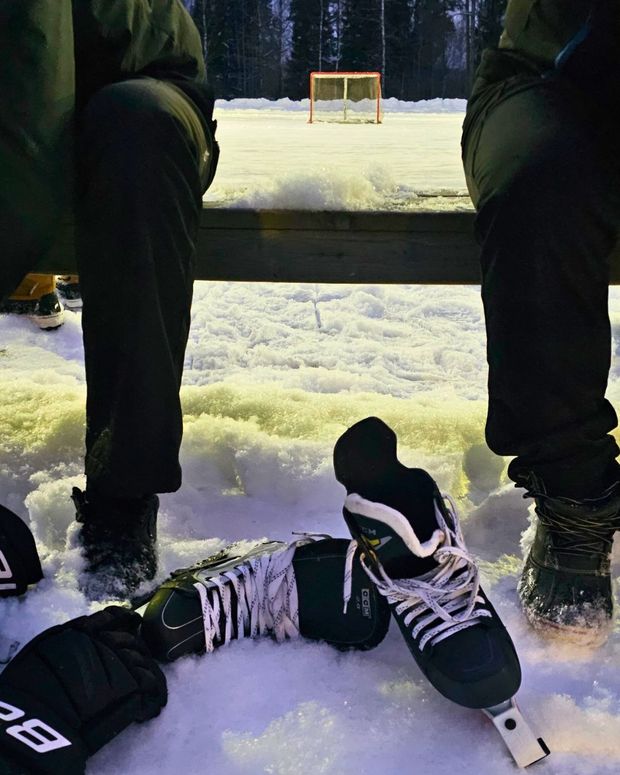 Talvinen ulkoj&auml;&auml;kiekkokentt&auml; h&auml;m&auml;r&auml;ss&auml;: etualalla lumessa lep&auml;&auml;v&auml;t luistimet ja hanskat penkin alla, taustalla n&auml;kyy punainen maali ja mets&auml;n reunaa.