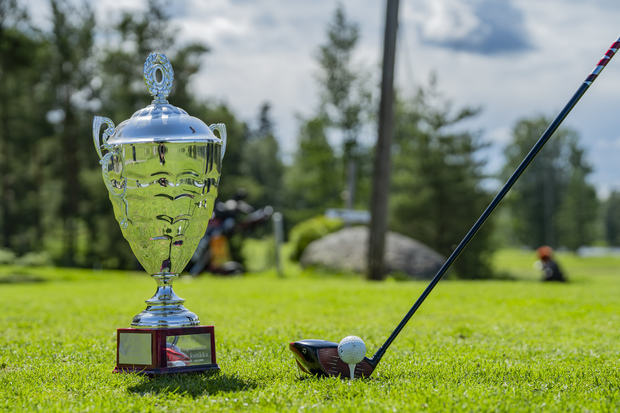 Hopeinen golfpalkintopokaali ja tiille asetettu golfpallo mailoineen nurmikolla, taustalla puustoa ja golfkent&auml;n maisemaa.