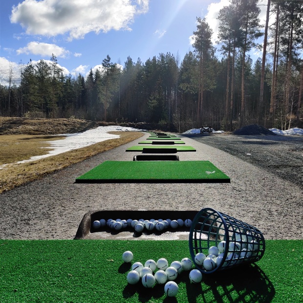 Golfpalloja kaatuneessa korissa ja ly&ouml;ntialustoja jonossa ulkorangella, taustalla mets&auml;inen maisema ja kev&auml;taurinko.