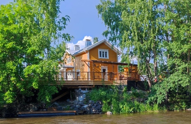 Keltainen koskisaunarakennus, ymp&auml;rill&auml; lehtipuita ja sininen taivas.